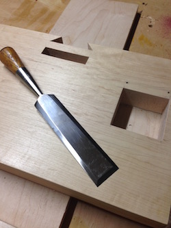 Large sharpened chisel on wood bench beside a large mortise and tenon joint in maple board.