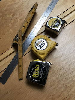 Magic of hand tools variety: three tape measures, folding ruler, and stainless steel rulers on woodworking bench.